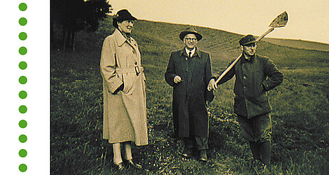 Three people are standing in a field: a woman in a long coat, a man in a suit and another man wearing a hat and carrying tools.