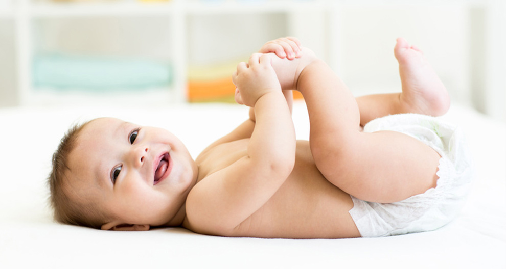 Een baby ligt op een wit oppervlak en houdt zijn voet vast.