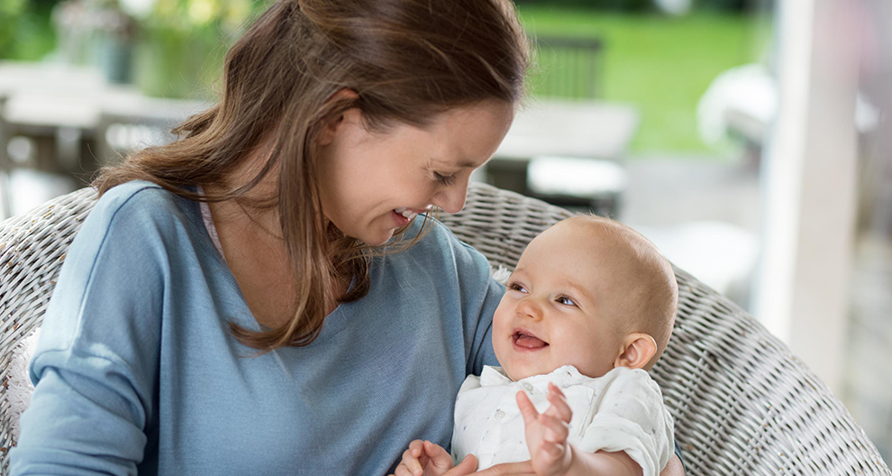Een vrouw in een blauw shirt houdt een baby vast in een witte outfit, beiden kijken elkaar aan met een patio op de achtergrond.