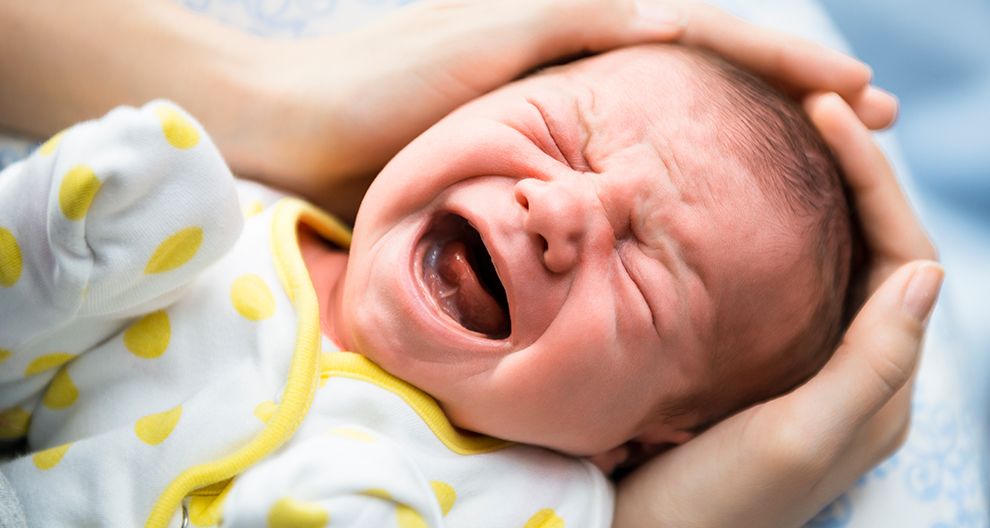 Een close-up van het gezicht van een huilende baby, vastgehouden door een hand, met een wit en geel gestippelde outfit.