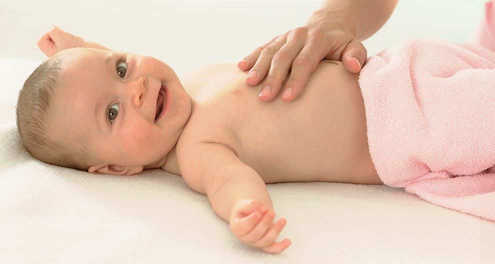 Een baby ligt op een wit oppervlak, gewikkeld in een roze handdoek, met een hand op de borst van de baby.