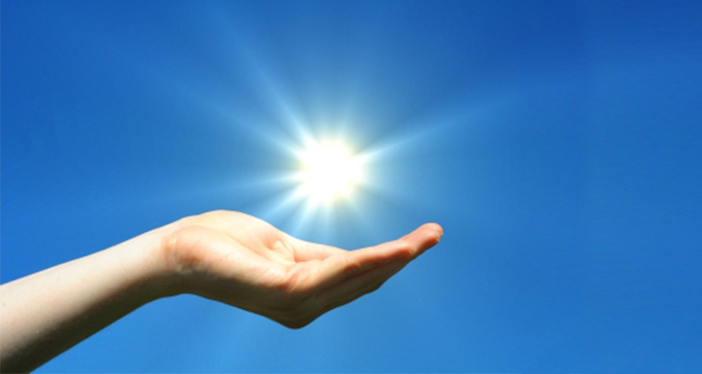 Een hand houdt een stralende zon tegen een helderblauwe lucht.