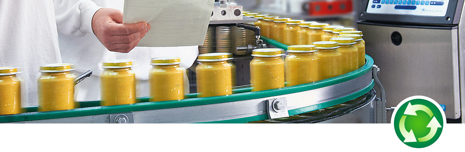 A worker in a white lab coat supervises a production line of yellow liquid-filled bottles moving on a conveyor belt.