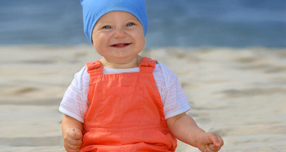 A baby wearing a blue hat and orange overalls sits on a sandy beach.