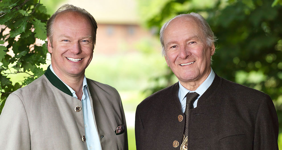 Two men, one in a light-colored jacket and the other in a dark-colored jacket, stand side by side in a garden setting.