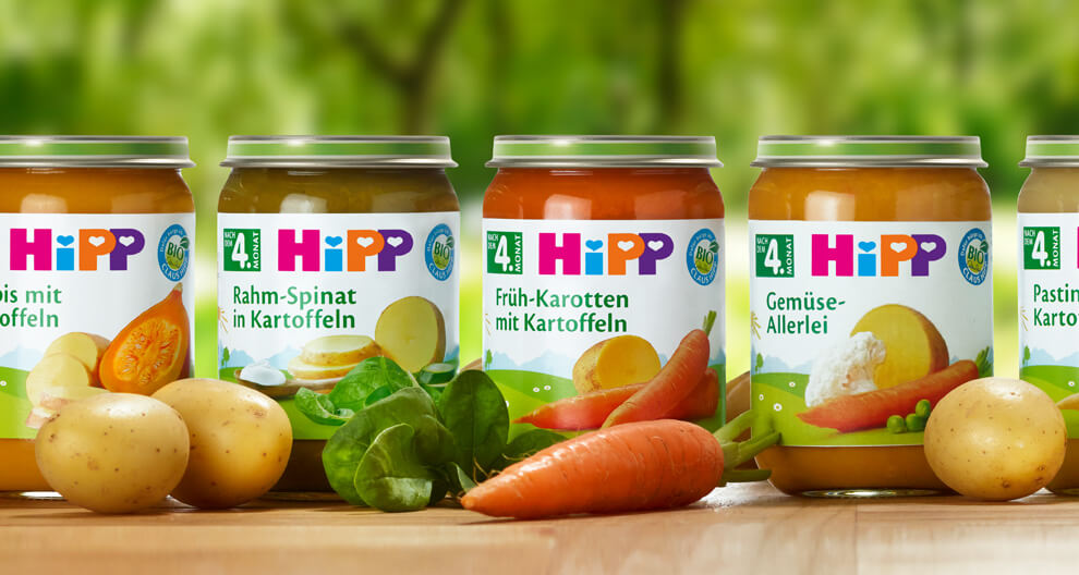 Four glass jars with labels displaying various food items are placed on a wooden surface, surrounded by raw vegetables.